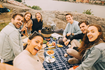 Portrait of cheerful friends enjoying picnic on field by lake during summer