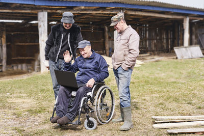 Happy senior man in a wheelchair in the yard , looking at a laptop. two friends are standing near