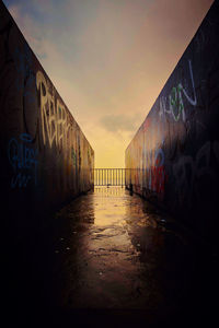 Wet walkway amidst graffiti wall against sky during sunset