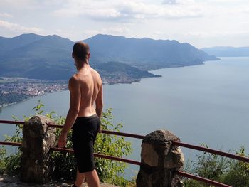 Rear view of shirtless man looking at sea against mountains