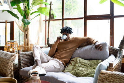 Young african american male drinking fresh coffee while sitting on comfortable sofa in light living room with potted plant at home