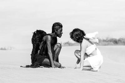 Side view of angel and devil kneeling on sand at beach against clear sky