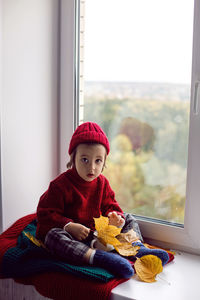 Boy a child in a red sweater and a knitted hat sits at the window on the windowsill