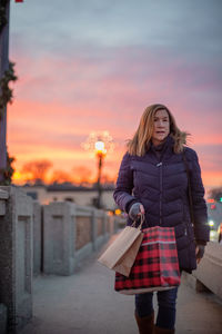 Woman looking away while walking on footpath against sky during sunset