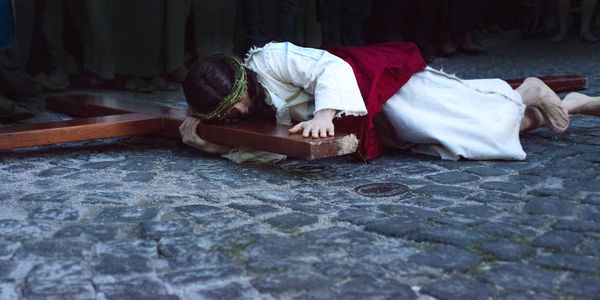 Man in jesus christ costume lying with wooden cross on footpath