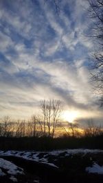 Bare trees on snow covered field against sky during sunset