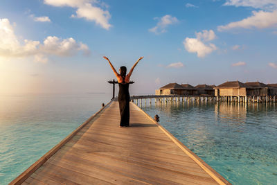 Rear view of woman with arms raised standing on pier over sea against sky