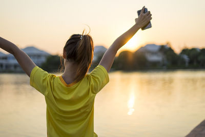 Rear view of woman standing by lake against sky during sunset