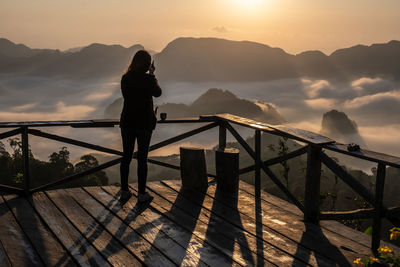 Woman standing on railing against sky during sunset