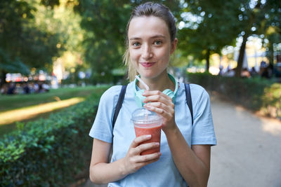 Joyful blonde teen female with cold drink spending summer day in park looking at camera
