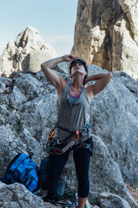 Young fit female climber in safety harness and helmet looking up while standing in rocky mountains and preparing for mountaineering looking up