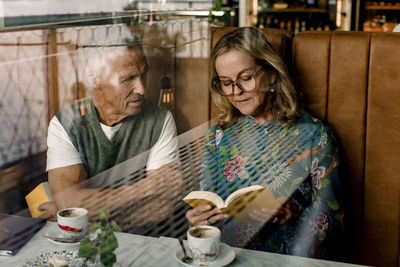 Senior woman reading book to male friend sitting with arms crossed in cafe seen through glass