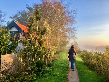 Rear view of a woman walking at sunrise on a small rural dike along a red-roofed farmhouse