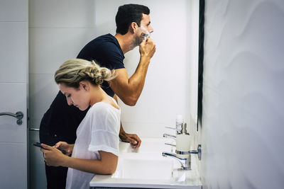 Man shaving with girlfriend using phone in bathroom