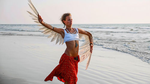 Young woman with costume wing standing at beach against sky