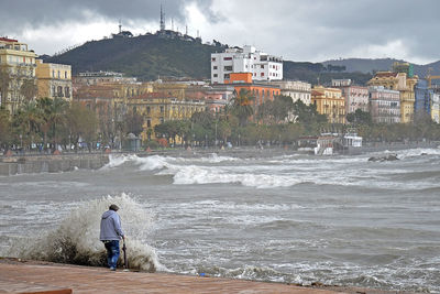 Rear view of man and buildings by sea against sky in city