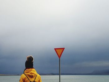 Rear view of woman in warm clothing standing by signboard at beach against cloudy sky