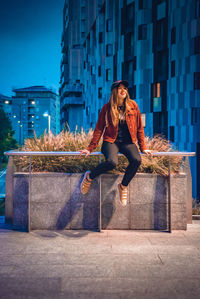 Full length of young woman sitting against buildings in city during dusk