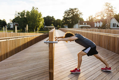 Side view of male athlete stretching legs while warming up during workout on wooden boardwalk n summer