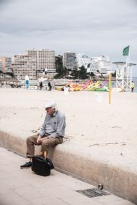 Man sitting at beach against sky in city