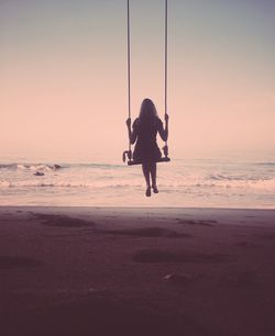 Full length rear view of women on swing at beach against clear sky