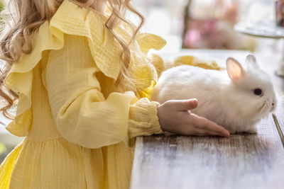 A little blonde girl in a yellow dress is sitting at a festive easter table with rabbits.