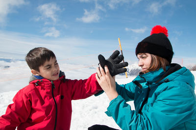 Mother helping son in wearing glove on snow against sky