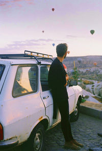 Side view of man standing by car against hot air balloons in sky