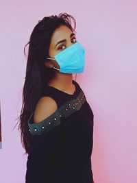 Portrait of young woman standing against pink background wearing surgical mask 