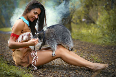 Young woman in traditional clothing playing with raccoon in forest