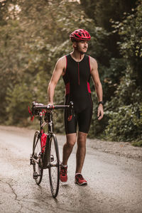 Full length of young man with bicycle walking on road against plants