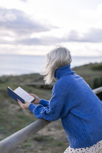 Side view of unrecognizable young female in blue sweater leaning on wooden fence and enjoying favorite poetry while spending time at seaside