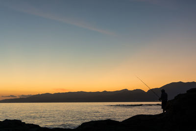 Silhouette man fishing in sea against sky during sunset