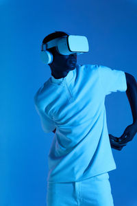 Cool young african american male in white outfit and futuristic vr goggles with headphones looking away while standing against blue background with hands behind back