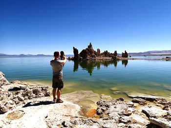 Full length rear view of man photographing sea using camera against clear blue sky