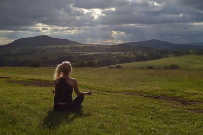 Woman is meditating in a mountain meadow in late afternoon with face turned to the sun behind clouds