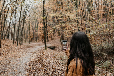 Young woman in yellow jacket taking photos with mobile phone in forest, autumn, fall.