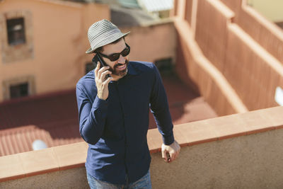 Attractive middle-aged man with hat and sunglasses talking to a smart phone standing on balcony.