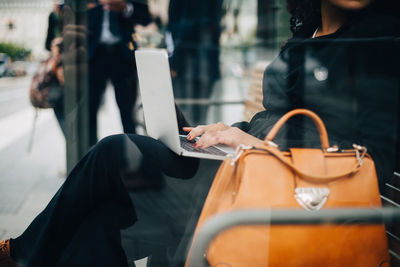 Midsection of businesswoman using laptop sitting at bus shelter seen from glass