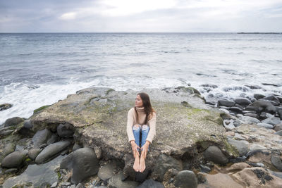 Woman sitting on rock at beach against sky