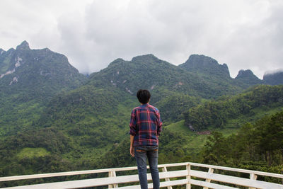 Rear view of man standing by railing against mountains and sky