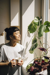 Smiling woman holding coffee cup while standing with eyes closed in sunlight at home