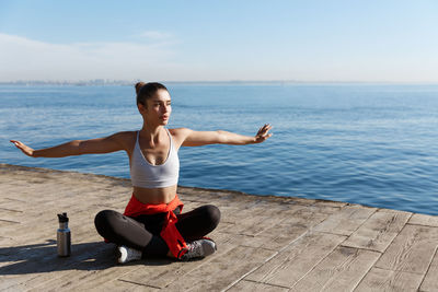 Full length of woman exercising while sitting against sea