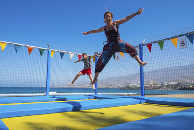 Cheerful woman jumping with son on trampoline against blue sky