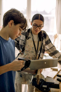 Male teenage student cutting wood while standing with female teacher during carpentry class