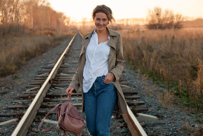 Young attractive lady in white shirt and beige raincoat walking along the railroad tracks 