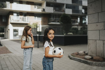 Girl holding ball while standing near sister on footpath