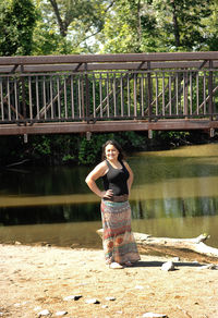Full length of smiling woman standing by lake against bridge and trees