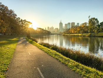 Diminishing perspective of empty road by river against clear sky in city during sunset