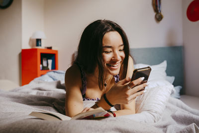 Happy young woman using smart phone while lying on bed at home